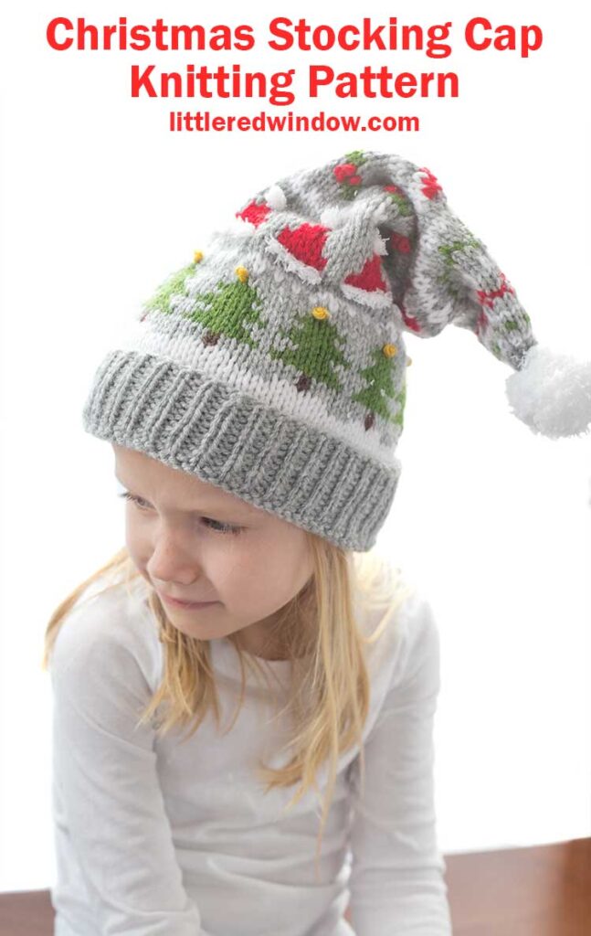 blond girl in white long sleeved shirt wearing a gray knit stocking cap decorated with patterns of Christmas trees Santa hats holly berries and candy cane stripe and a white pom pom on top looking down and off to her left