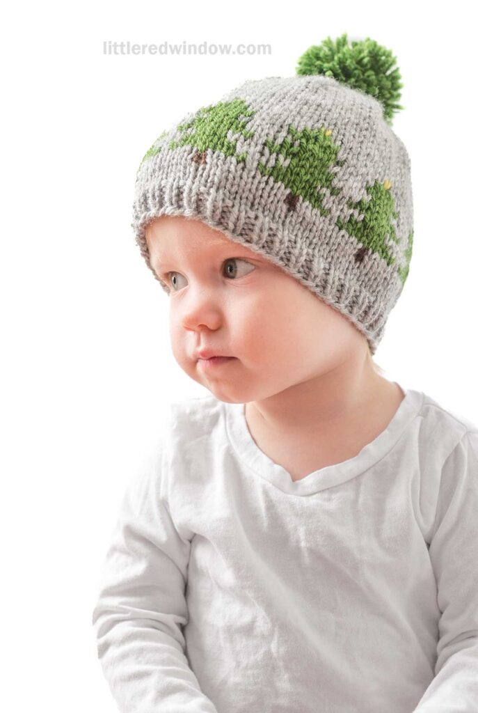 A baby wearing a white shirt and a charming Christmas Tree Farm Hat with green and gray patterns, complete with a playful green pom-pom on top, sits against a white background. The baby looks to the side.