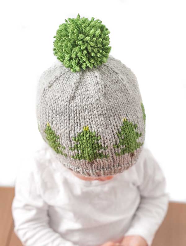 A child in a gray Christmas Tree Farm hat, featuring green pom-poms and tree designs, sits facing downward against a white background. The child's clothing is pure white, creating a serene and cozy winter scene.