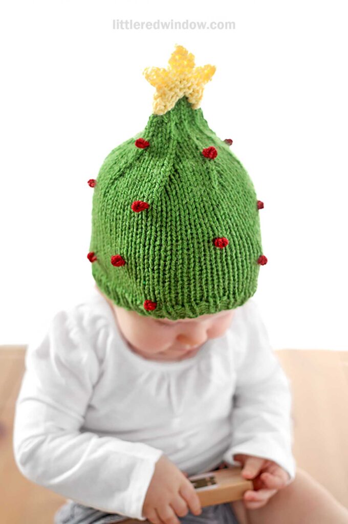 A baby is sitting and looking down while holding a small toy, wearing a charming Christmas tree hat knit in green with a yellow star on top and red decorations.