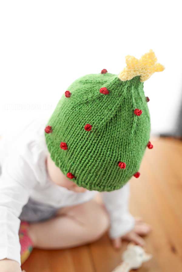 A baby sits on a wooden floor, wearing an adorable knitted Christmas tree hat. The hat is decorated with small red ornaments and crowned with a charming yellow star.