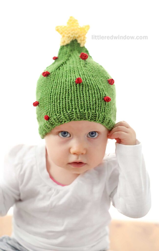 A baby wearing a whimsical Christmas tree hat, adorned with a yellow star and red ornaments, is dressed in a white shirt and seems to be adjusting the festive green knitted creation.