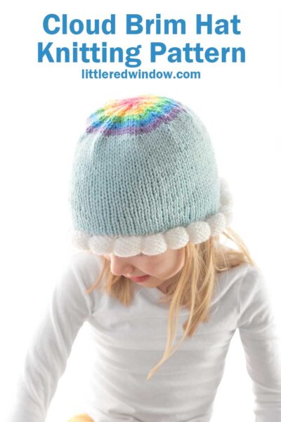 blond girl wearing light blue knit hat with bubble knit white cloud brim and a rainbow decrease section on the top in front of a white background looking down to the left