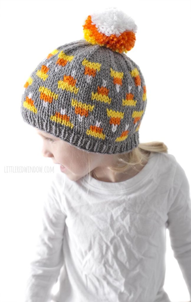 little girl in white shirt looking to the left and wearing gray knit hat with candy corn pattern on it and candy corn colored pom pom on top