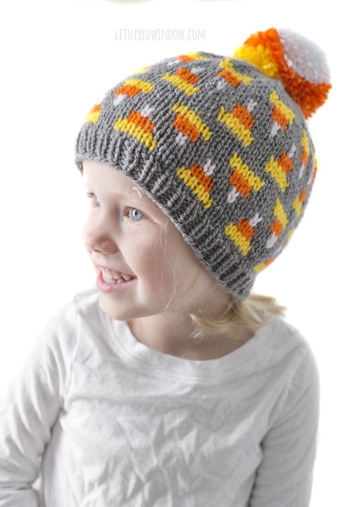 little girl in white shirt looking off to the left and wearing gray knit hat with candy corn pattern on it and candy corn colored pom pom on top