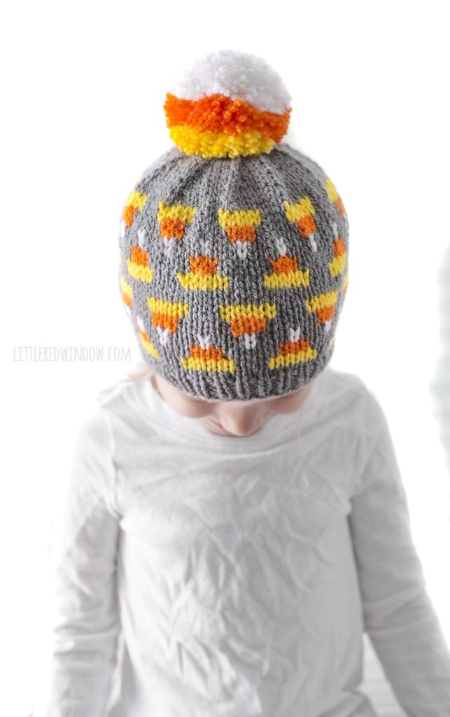 little girl in white shirt looking down and wearing gray knit hat with candy corn pattern on it and candy corn colored pom pom on top