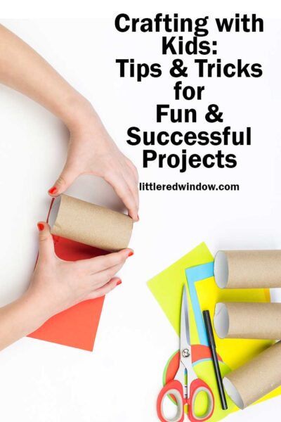 a childs hands holding a cardboard tube over orange paper next to various craft supplies on a white tabletop