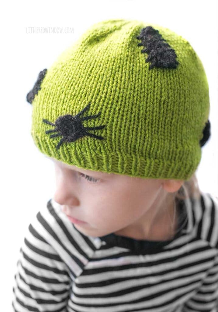 child in black and white striped shirt wearing an acid green knit hat with black knit spiders and bugs on it leaning forward and looking off to the left in front of a white background