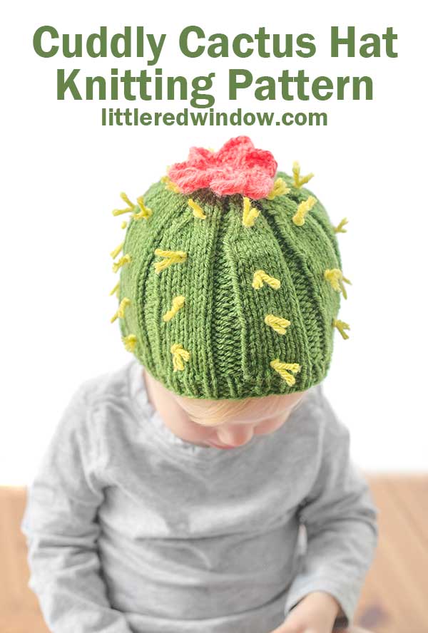 Child in gray shirt on a wood table wearing a medium green ribbed knit hat with light green yarn spikes and a pink cactus flower on top to make a cactus hat looking down at their lap