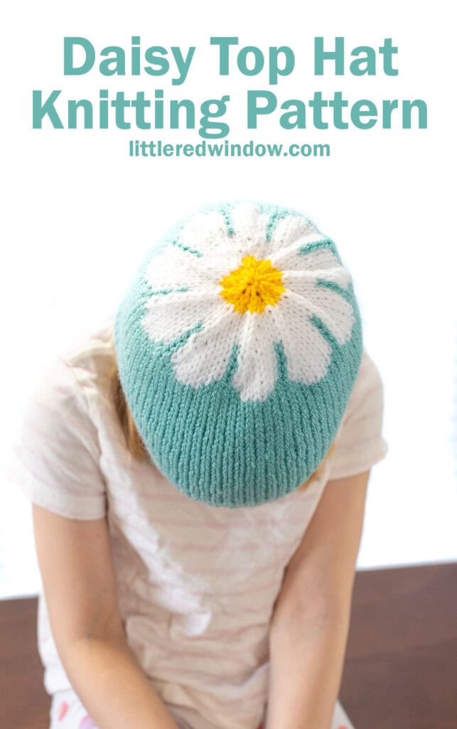 child leaning forward to show the top of a blue knit hat with a white and yellow daisy pattern centered on the top of it
