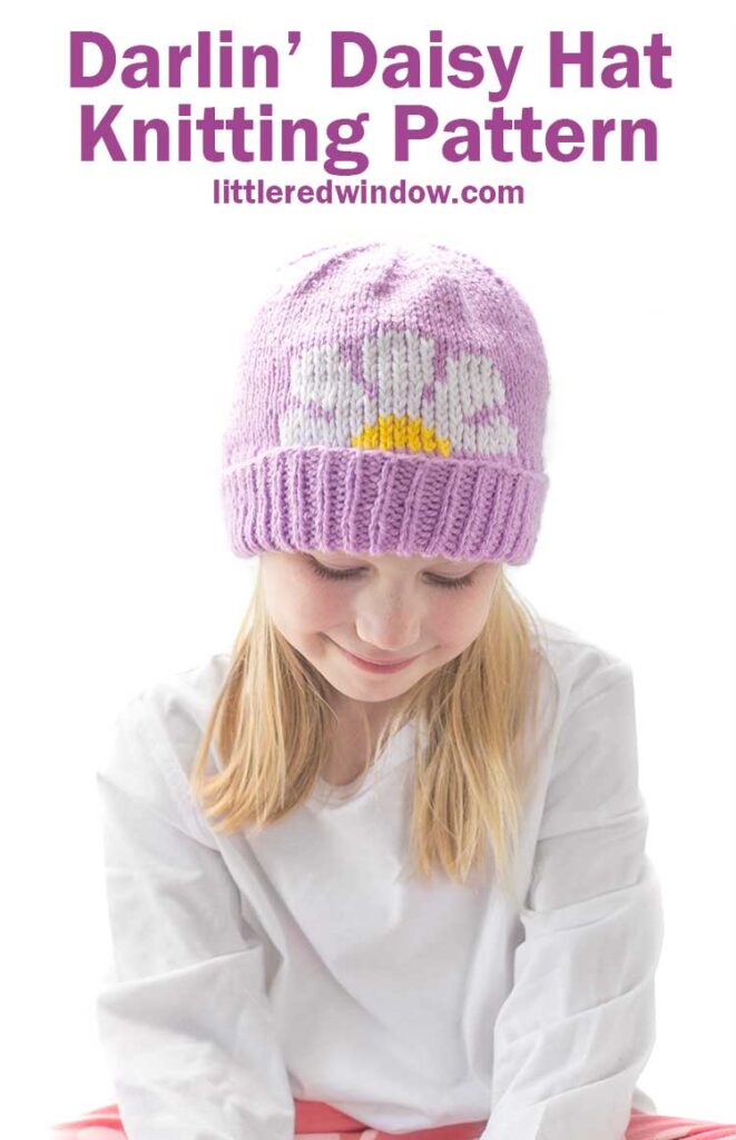 Smiling blonde girl looking down while wearing a white shirt and a lavender knit hat with thick folded brim that has a white and yellow daisy embroidered on the front peeking halfway over the brim