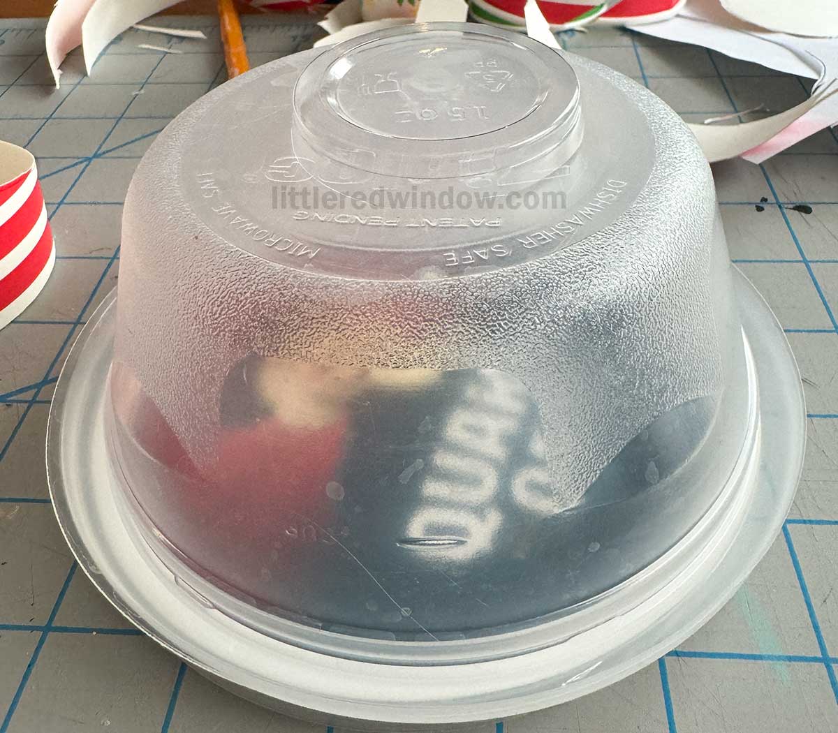 A plastic bowl placed upside down on a circular oats lid.. The background includes a grid-patterned surface and scattered paper strips.