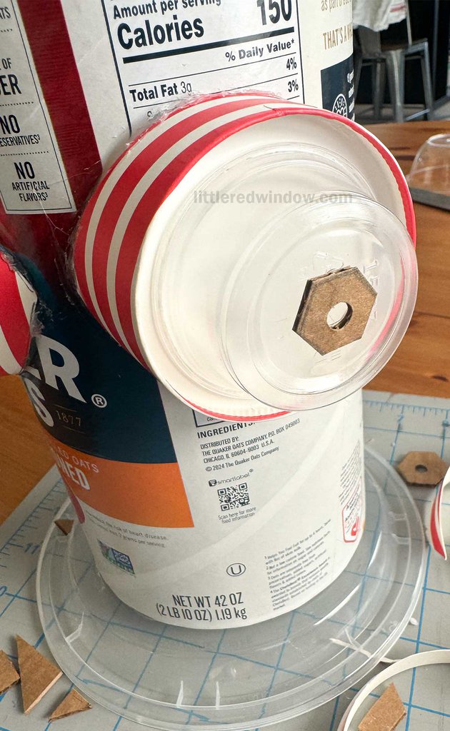 A craft project showing an oatmeal container being transformed into a DIY fire hydrant. The container has a plastic lid, red and white striped tape, and a cardboard circle with a nut in the center on the front. Craft materials are scattered around.