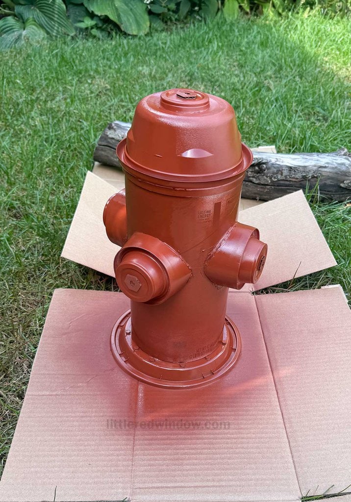 A fire hydrant is sprayed with a fresh coat of orange-red primer, placed on two pieces of cardboard on a grassy lawn. A log can be seen in the background.
