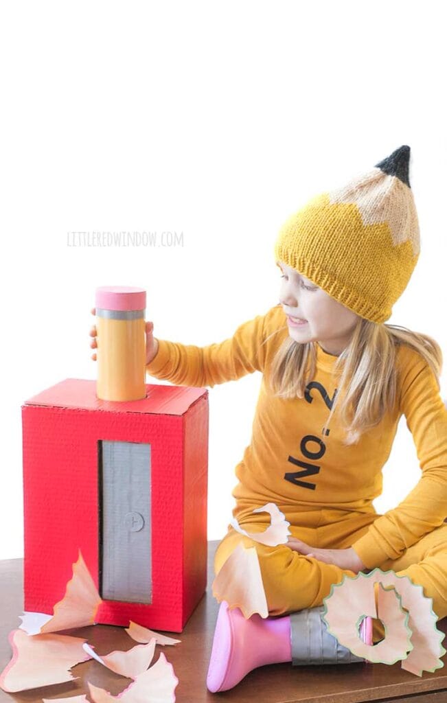 girl sitting with crossed legs wearing a pencil costume on a wood table next to a large red pencil sharpener with a pencil sticking out of the top and large pencil shavings all around her reaching over to grab the large pretend pencil