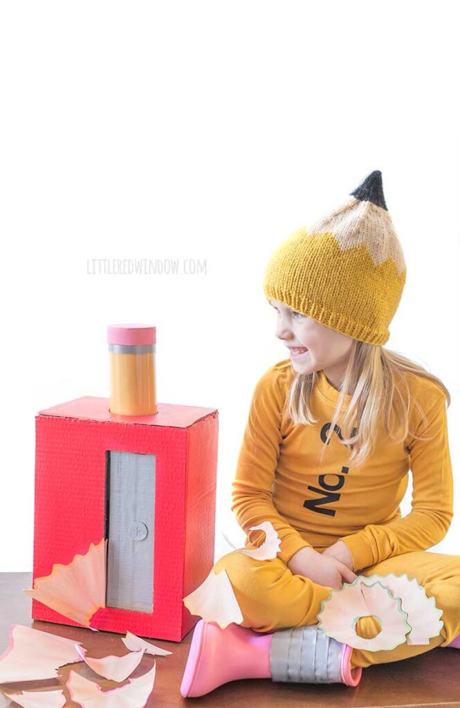 girl sitting with crossed legs wearing a pencil costume on a wood table next to a large red pencil sharpener with a pencil sticking out of the top and large pencil shavings all around her looking off to the left