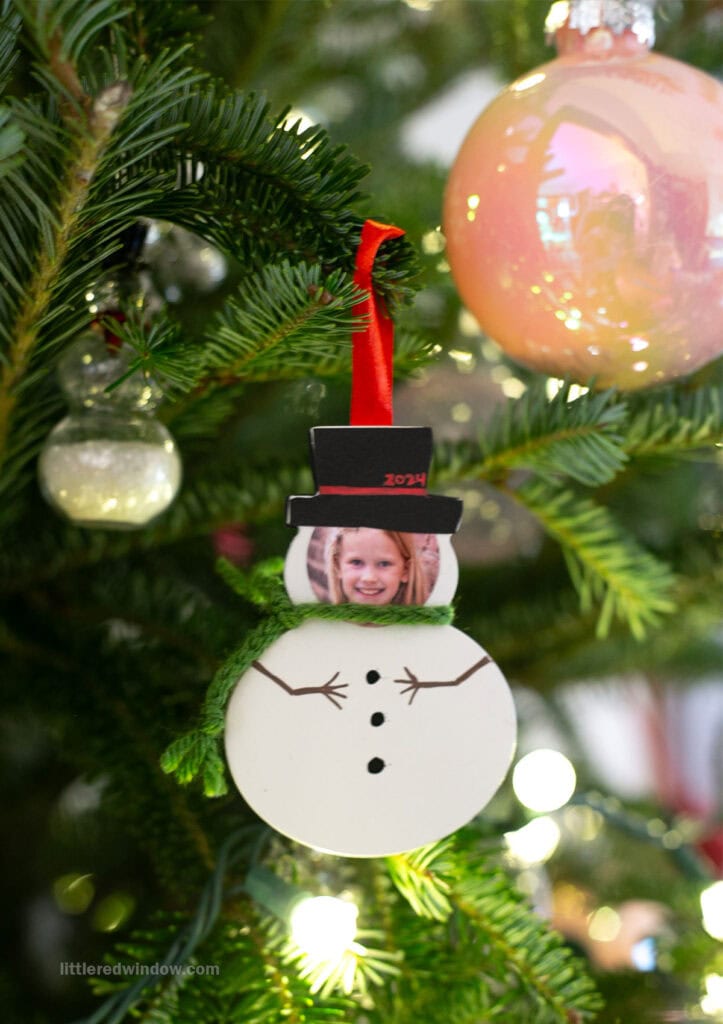 A snowman ornament with a photo of a smiling child as its face hangs on a Christmas tree. The ornament has a red ribbon, a black hat labeled