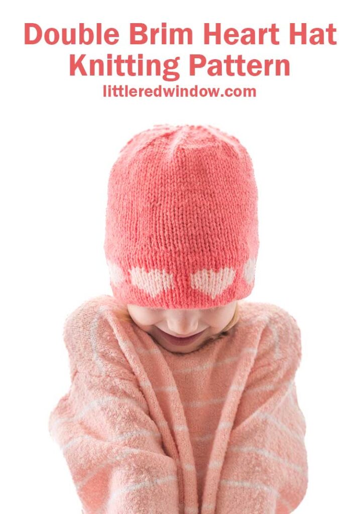 A child wears a pink double brim heart hat with white hearts and a matching sweater, playfully covering their mouth. Above them, the text reads