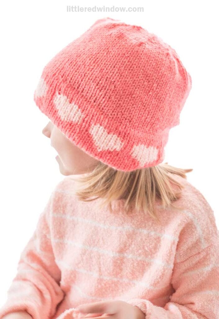 A child wearing a pink double brim heart hat with white heart patterns along the bottom edge complements their light pink sweater. The child's sideways glance stands out against the plain, bright background.