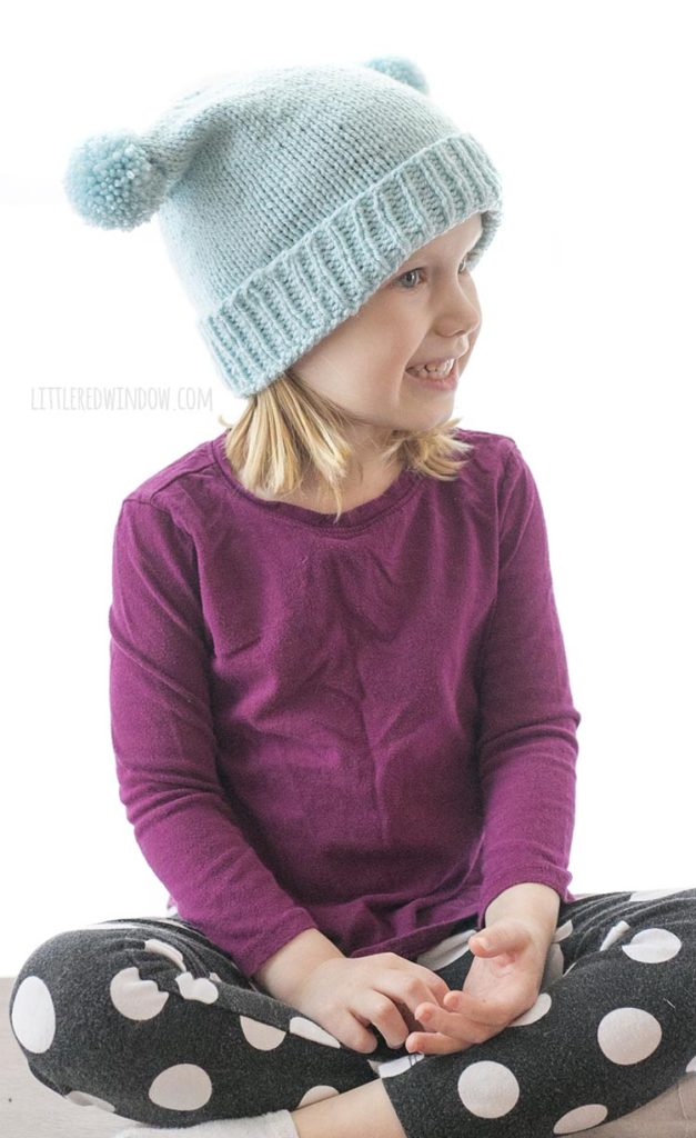 little girl wearing light blue hat with folded brim and two pom poms on the sides