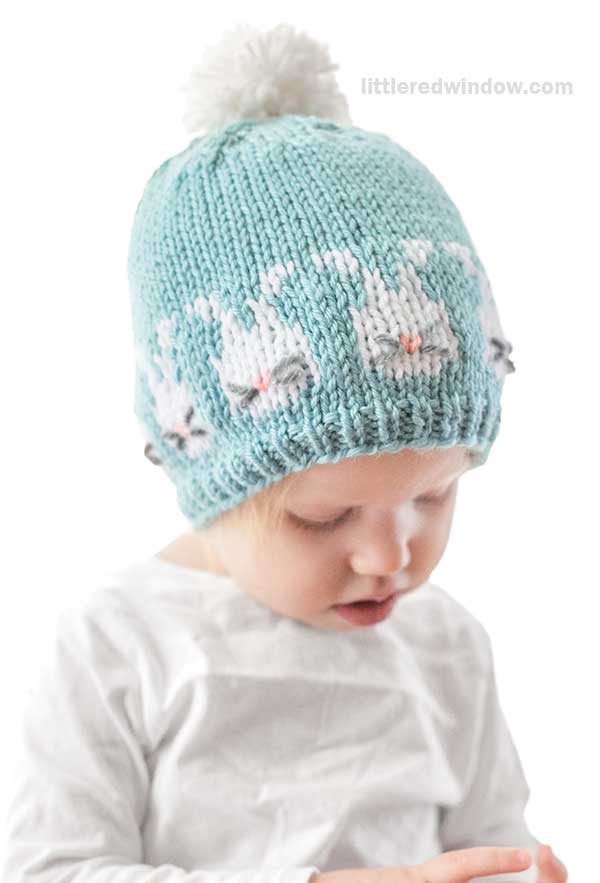 A young child wears a light blue knitted hat resembling Easter bunnies, complete with white pom-pom and cute animal ears. The child is looking down while dressed in a white shirt against a plain white background.