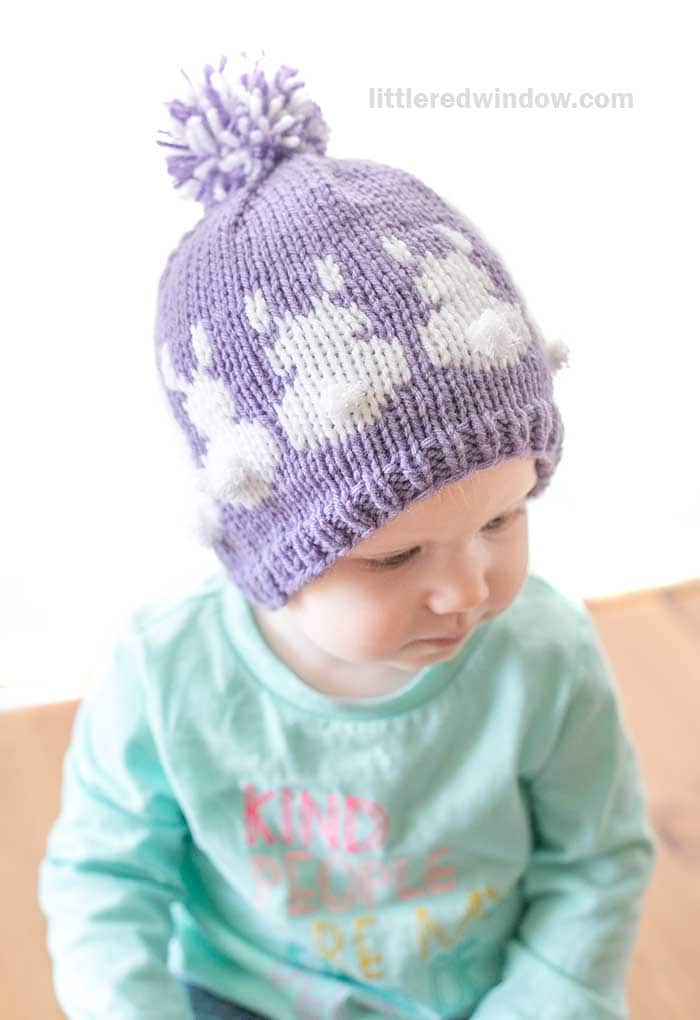 A child wearing an Easter Bunny hat, a charming purple knit adorned with white bunny patterns and a pom-pom, sits indoors. The young one also sports a light blue shirt featuring colorful text.