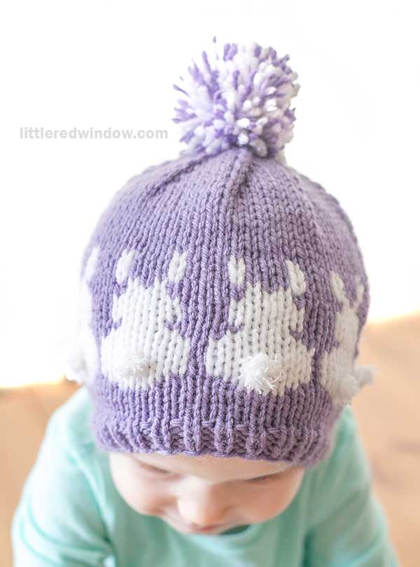 A baby wearing a purple knitted hat adorned with bunny patterns and a pom-pom resembles an Easter Bunny Hat. Dressed in a light blue outfit, the softly blurred background frames the scene, while the