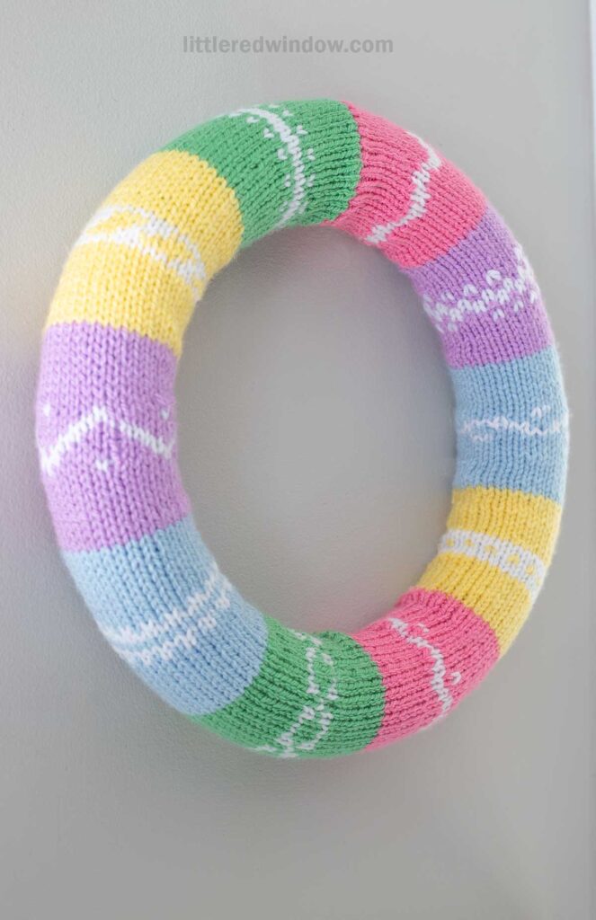 A colorful knitted wreath hangs on a wall. It features horizontal stripes in pastel colors: green, pink, yellow, blue, and purple, each adorned with white patterns resembling Easter eggs.