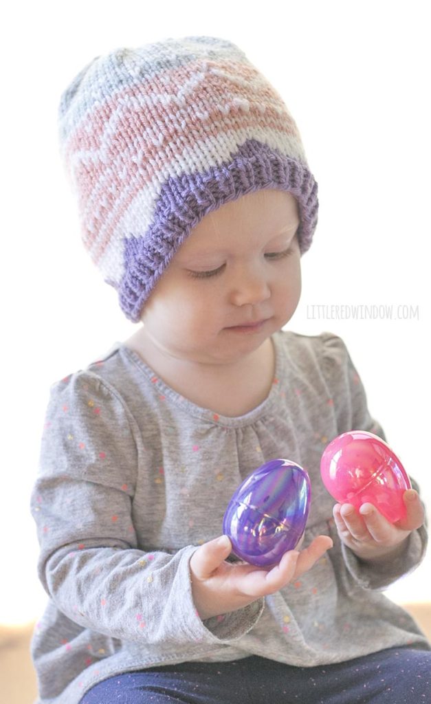 baby girl wearing knit easter egg hat and examining two pink and purple easter eggs in her hands