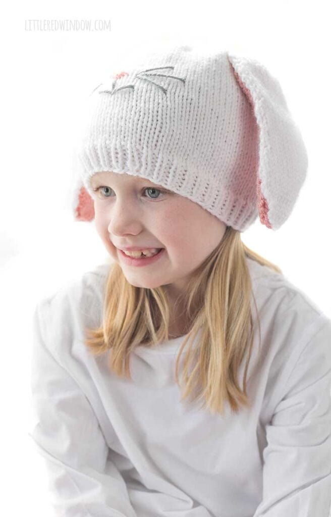 Smiling girl in white shirt wearing a white knit hat with long floppy white bunny ears with pink lining and a cute bunny face on the front looking down and to the left