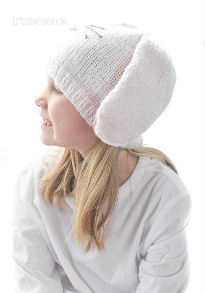 Smiling girl in white shirt wearing a white knit hat with long floppy white bunny ears with pink lining and a cute bunny face on the front looking off to the left