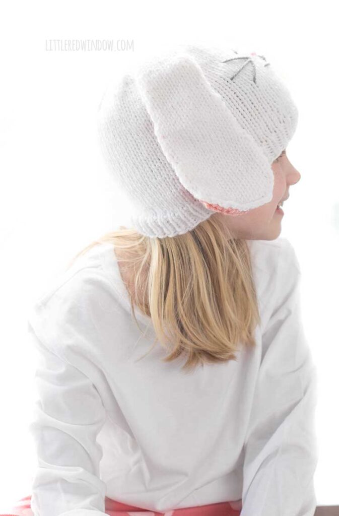 Smiling girl in white shirt wearing a white knit hat with long floppy white bunny ears with pink lining and a cute bunny face on the front looking off to the right