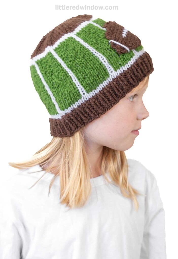 A child wears a football hat designed to look like a football field, featuring green, white, and brown colors with a small football detail on the side. The child is facing to the right.