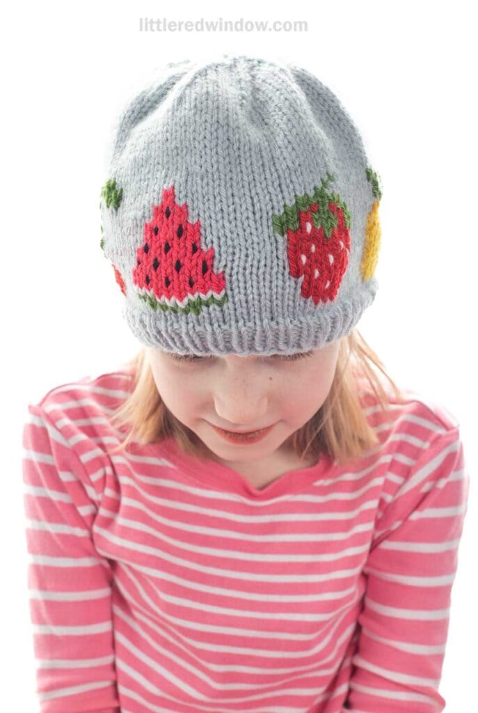 A young girl wears a fruit salad hat—a gray knit cap adorned with watermelon, strawberry, and pineapple—paired with a pink and white striped shirt. She looks down, smiling slightly.