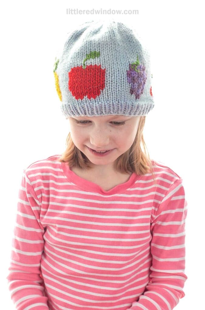 A young girl wearing a pink and white striped shirt and a light blue knit fruit salad hat decorated with colorful designs, including an apple and grapes, looks down and smiles.