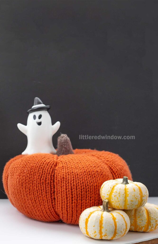 A small plastic ghost wearing a hat stands on a large knitted orange pumpkin. Three small white and yellow pumpkins are placed in front of it against a dark background. The text