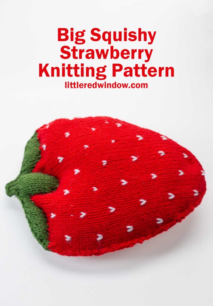A large, plush knitted pillow shaped like a strawberry with green leaves and white seed details. Red text above reads: