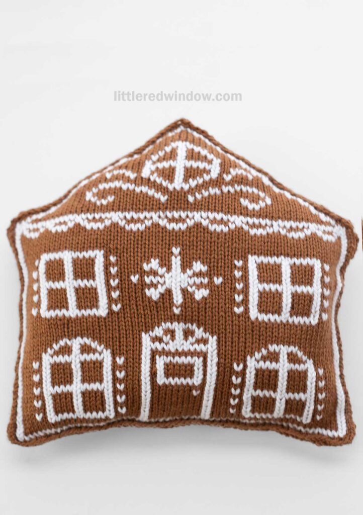 A knitted pillow shaped like a gingerbread house, featuring white decorative patterns resembling windows, a door, and snowflakes on a brown background.