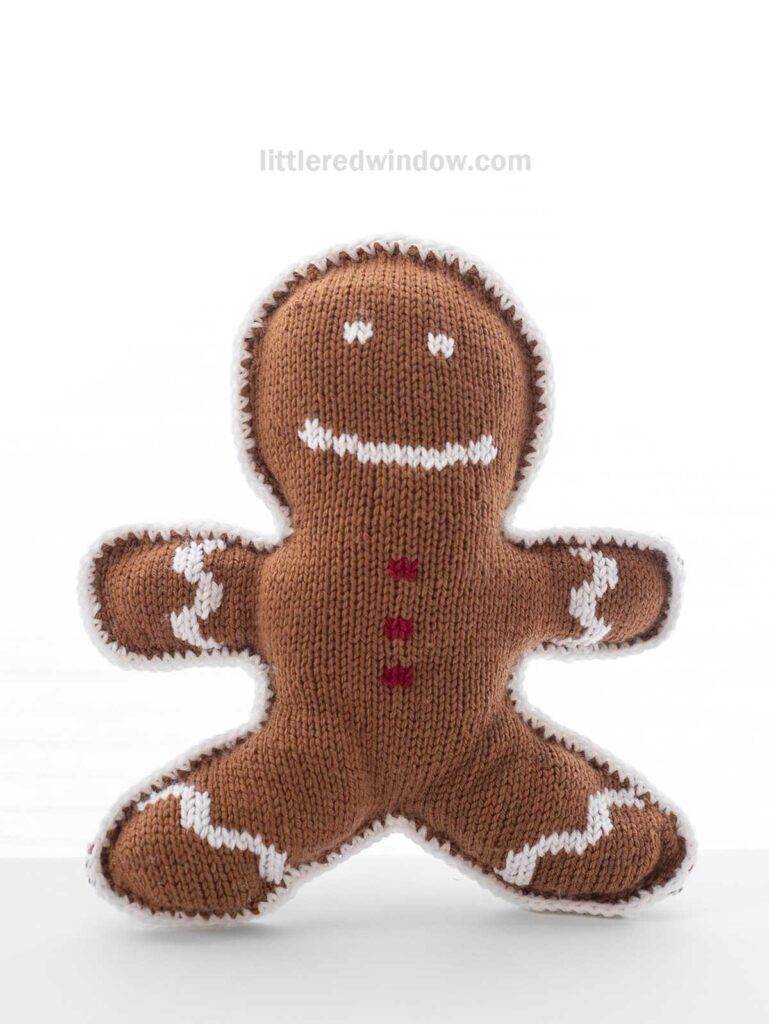 A knitted brown plush gingerbread man with white trim and red buttons, smiling against a plain white background.