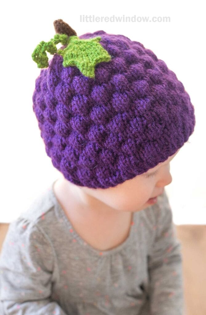 A baby wearing a grape hat, designed to look like a purple knitted grape with a green leaf and vine on top, is dressed in a light gray outfit with small colorful dots and is looking slightly downward.