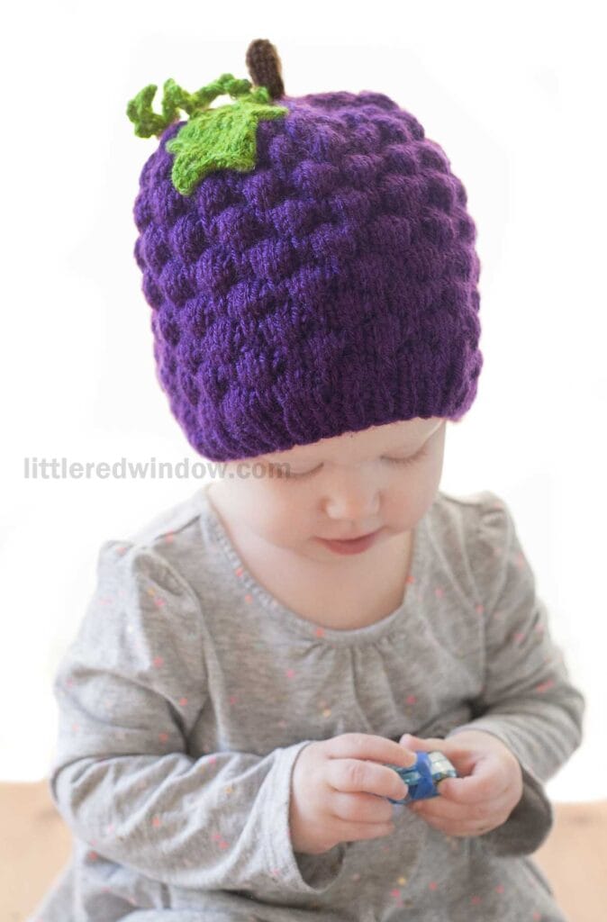 A young child wearing a grape hat resembling a cluster of grapes with green leaves and a brown stem looks down while holding a small object. The image features a watermark reading