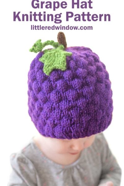 A baby is wearing a purple knitted grape hat, complete with a green leaf and stem on top. The text above reads, "Grape Hat Knitting Pattern," with "littleredwindow.com" underneath. The baby is dressed in a grey patterned outfit.