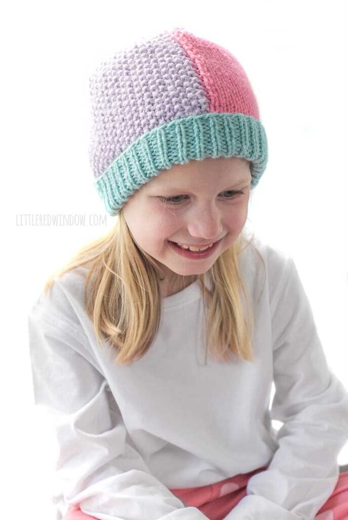 Smiling girl in white shirt and pink pants wearing a knit hat that has a light teal ribbed brim and is half pink stockinette stitch on the right and half light purple seed stitch on the left