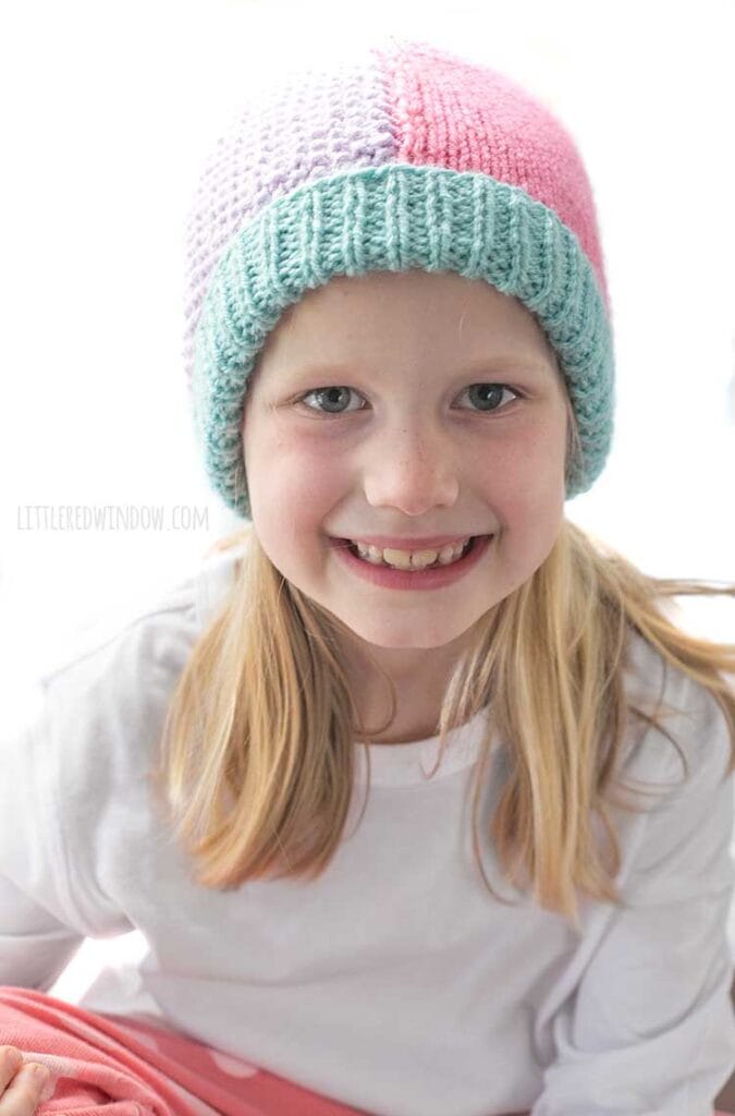 closeup of Smiling girl in white shirt and pink pants wearing a knit hat that has a light teal ribbed brim and is half pink stockinette stitch on the right and half light purple seed stitch on the left