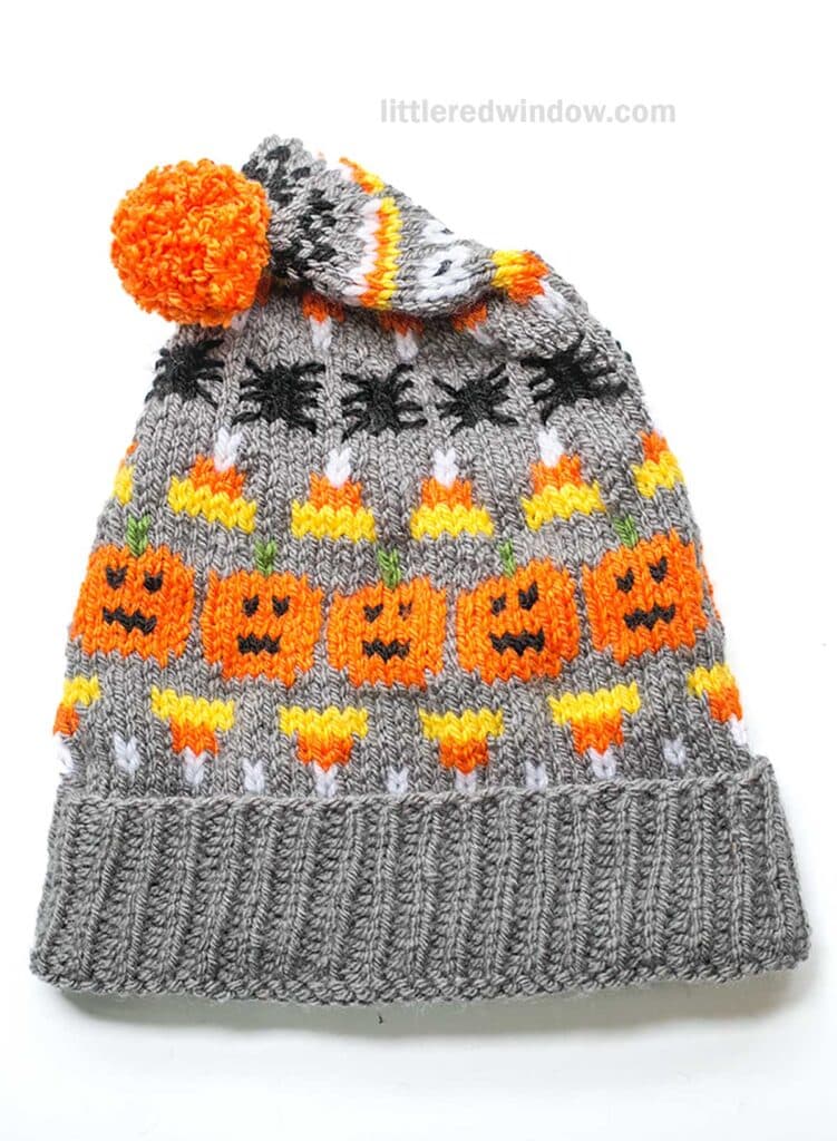 A knitted gray Halloween stocking cap features designs of orange pumpkins, black spiders, and yellow candy corn. The hat is topped with an orange pom-pom.
