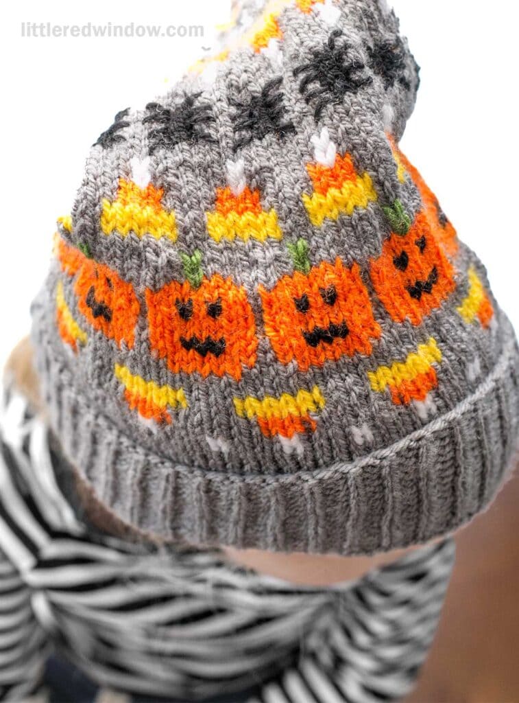 A young person wearing a gray Halloween stocking cap adorned with smiling orange pumpkins and yellow candy corn and spiders The person is also dressed in a black and white striped outfit, creating a festive look. The background is white and out of focus.