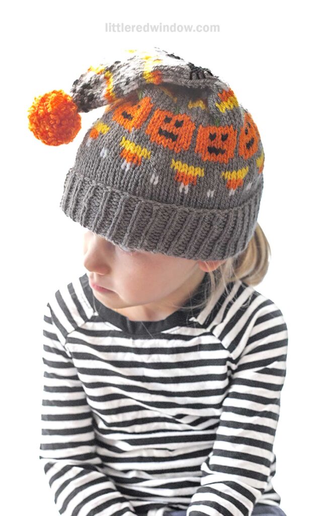 A child wearing a black and white striped shirt and a knitted gray beanie with orange and yellow Halloween patterns with an orange pom-pom on top, looks to the side. The background is white.
