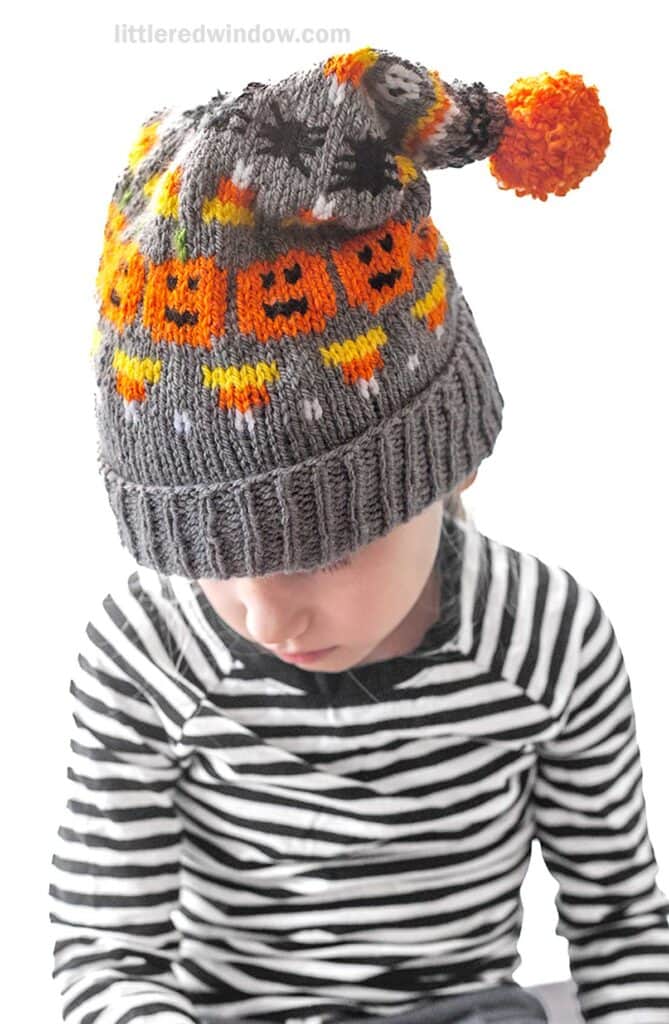 A child wearing a Halloween stocking cap with orange jack-o'-lantern, candy corn and spider patterns and a pom-pom on top. Dressed in a black and white striped shirt, the child is looking downward.