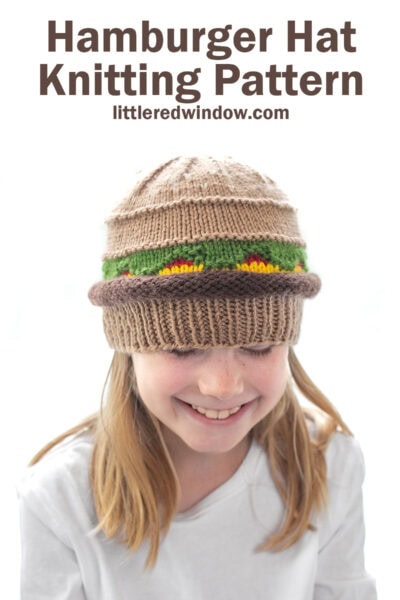 smiling girl in a white shirt wearing a knit hat that looks like a hamburger with beef, lettuce, ketchup and cheese layers looking down at their lap