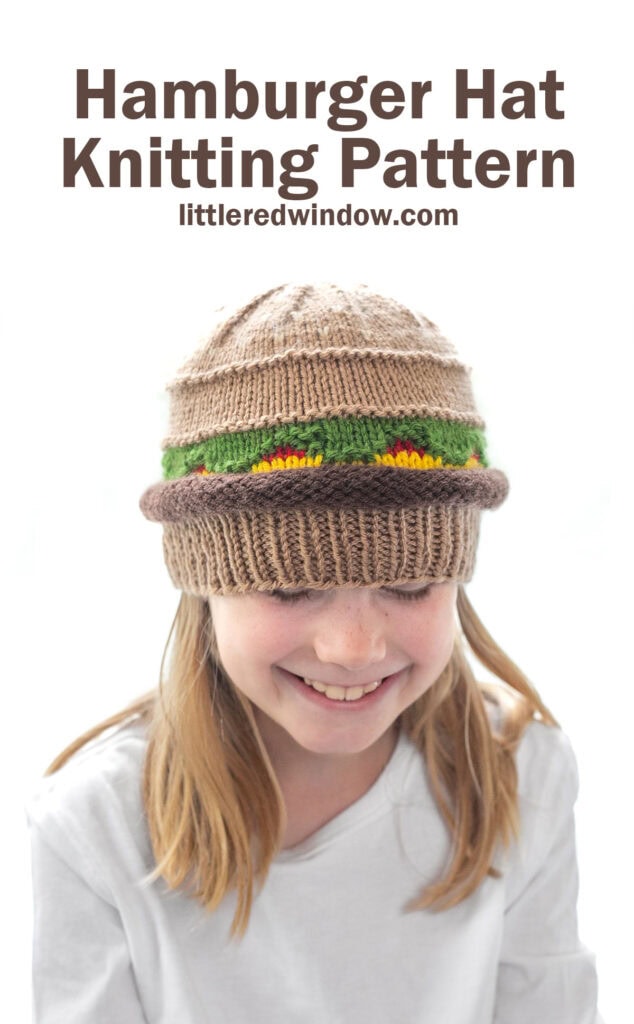 smiling girl in a white shirt wearing a knit hat that looks like a hamburger with beef, lettuce, ketchup and cheese layers looking down at their lap